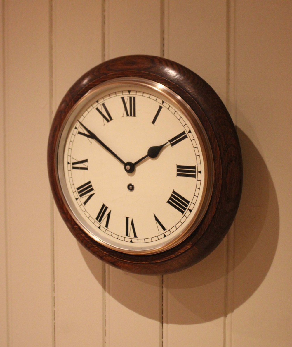 A circular early 20th-century wall clock featuring a moulded dark oak frame, a brass bezel, and a white dial face with black Roman numerals. View VI