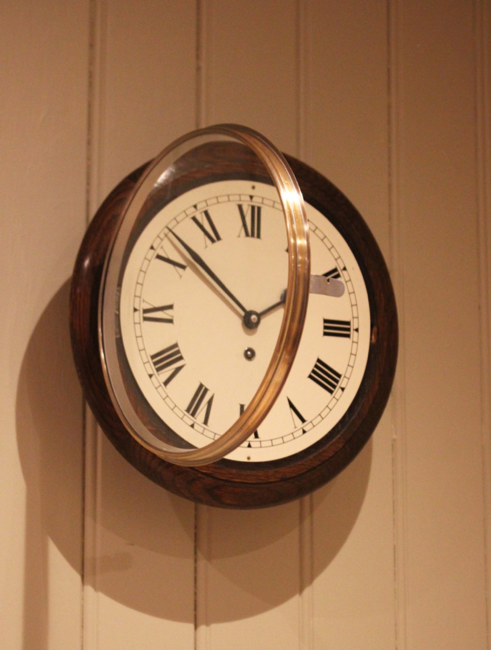 A circular early 20th-century wall clock featuring a moulded dark oak frame, a brass bezel, and a white dial face with black Roman numerals. View VIII