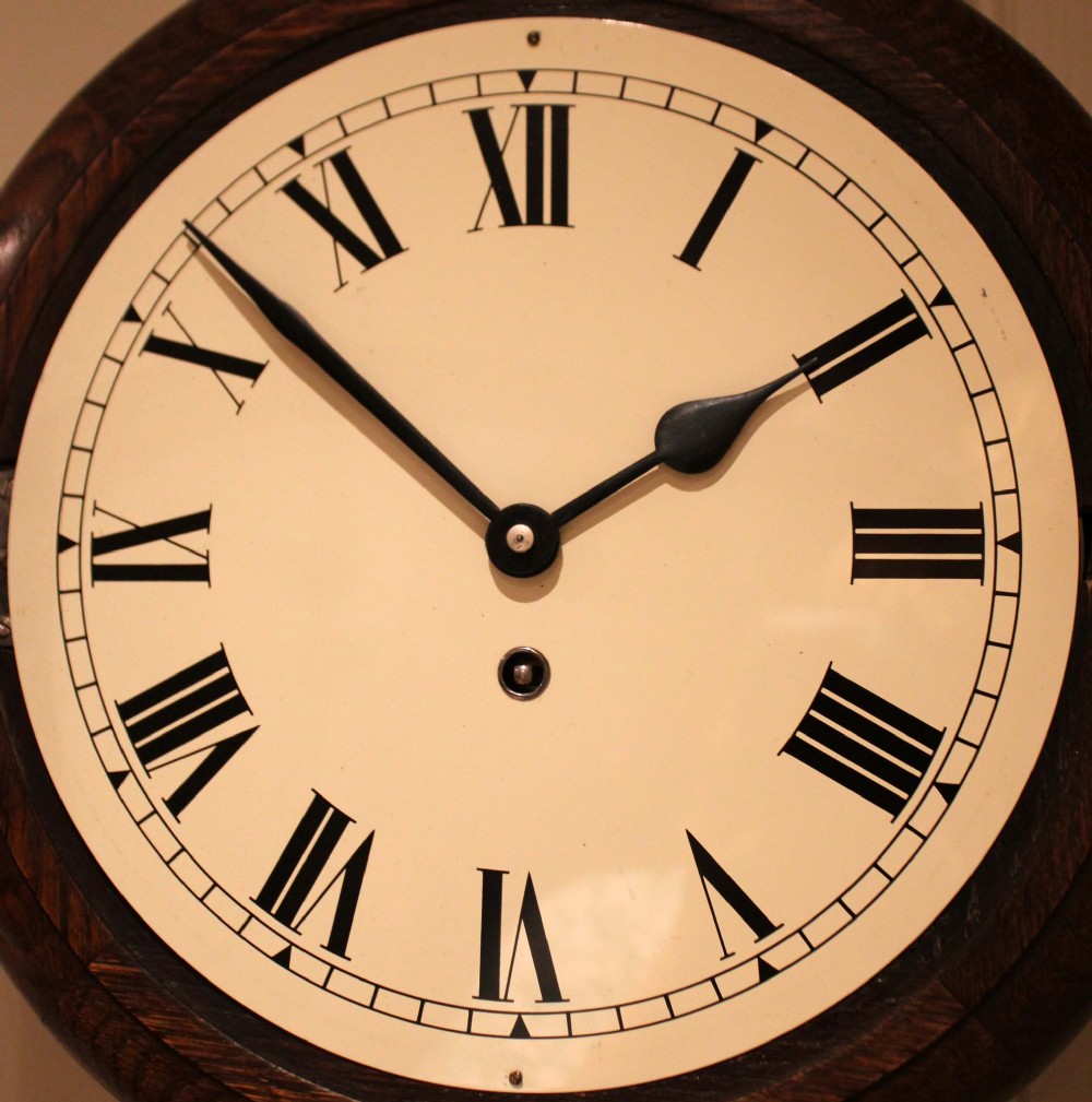 A circular early 20th-century wall clock featuring a moulded dark oak frame, a brass bezel, and a white dial face with black Roman numerals. View X