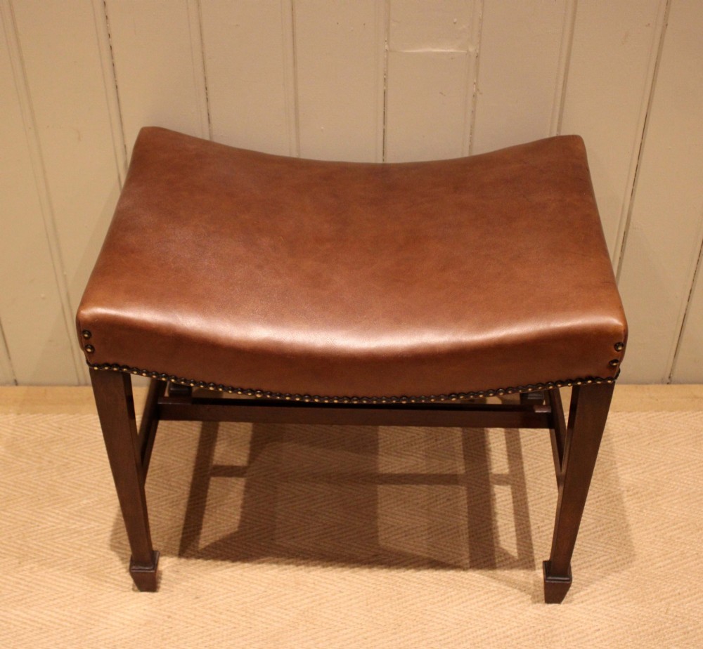 An Edwardian mahogany stool featuring a concave tan leather seat trimmed with brass studs and supported by square tapered legs with stretchers. View II