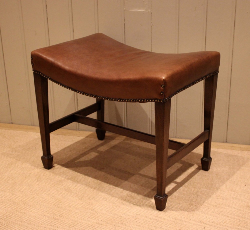 An Edwardian mahogany stool featuring a concave tan leather seat trimmed with brass studs and supported by square tapered legs with stretchers. View V