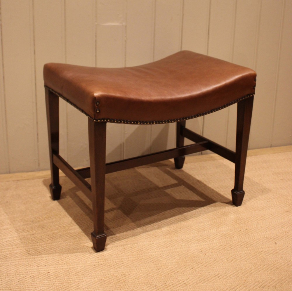 An Edwardian mahogany stool featuring a concave tan leather seat trimmed with brass studs and supported by square tapered legs with stretchers. View VI