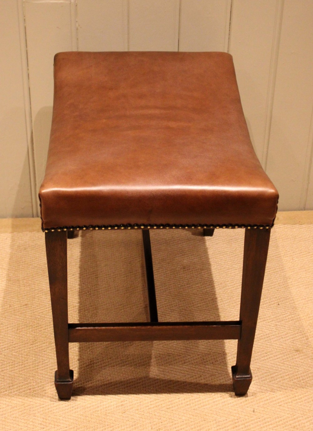 An Edwardian mahogany stool featuring a concave tan leather seat trimmed with brass studs and supported by square tapered legs with stretchers. View VII