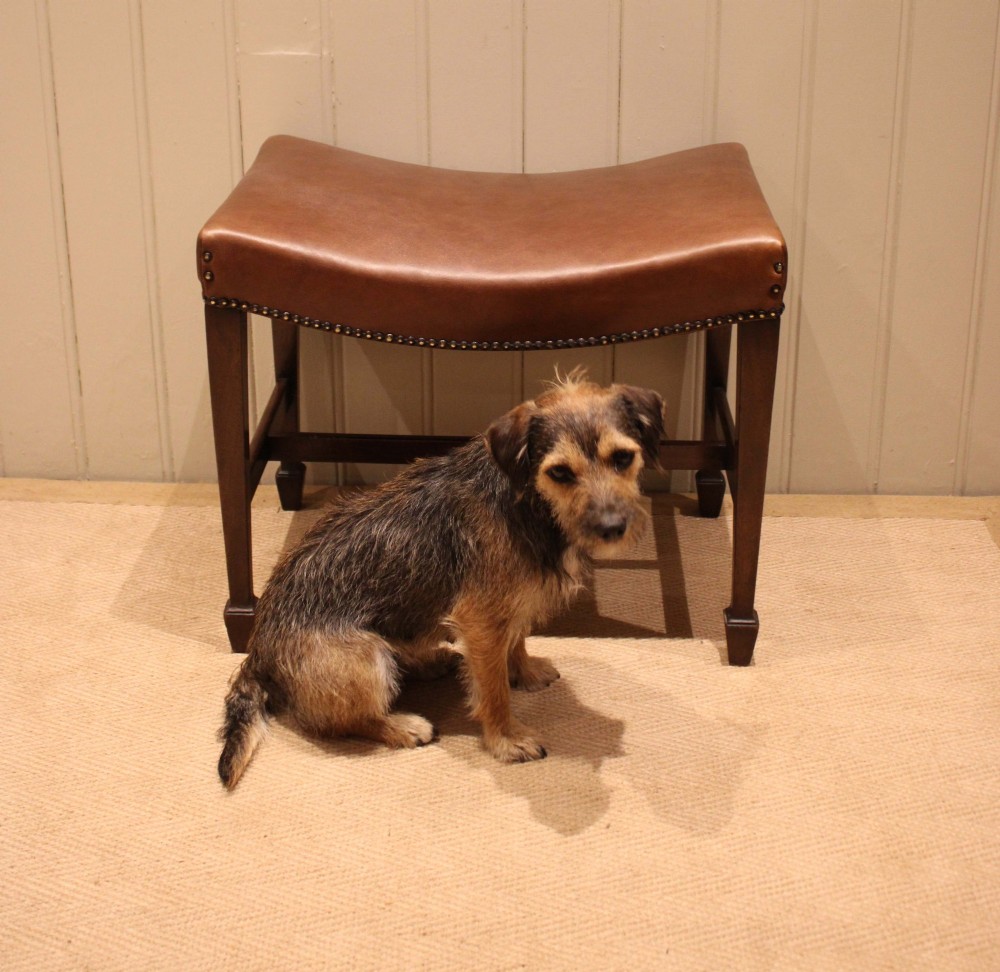 An Edwardian mahogany stool featuring a concave tan leather seat trimmed with brass studs and supported by square tapered legs with stretchers. View IX