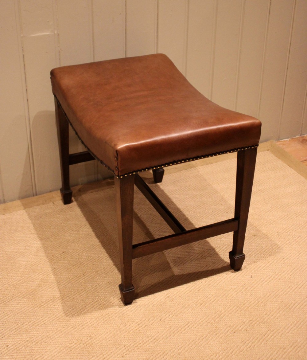 An Edwardian mahogany stool featuring a concave tan leather seat trimmed with brass studs and supported by square tapered legs with stretchers. View X