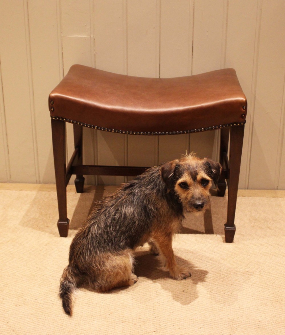 An Edwardian mahogany stool featuring a concave tan leather seat trimmed with brass studs and supported by square tapered legs with stretchers. View XI