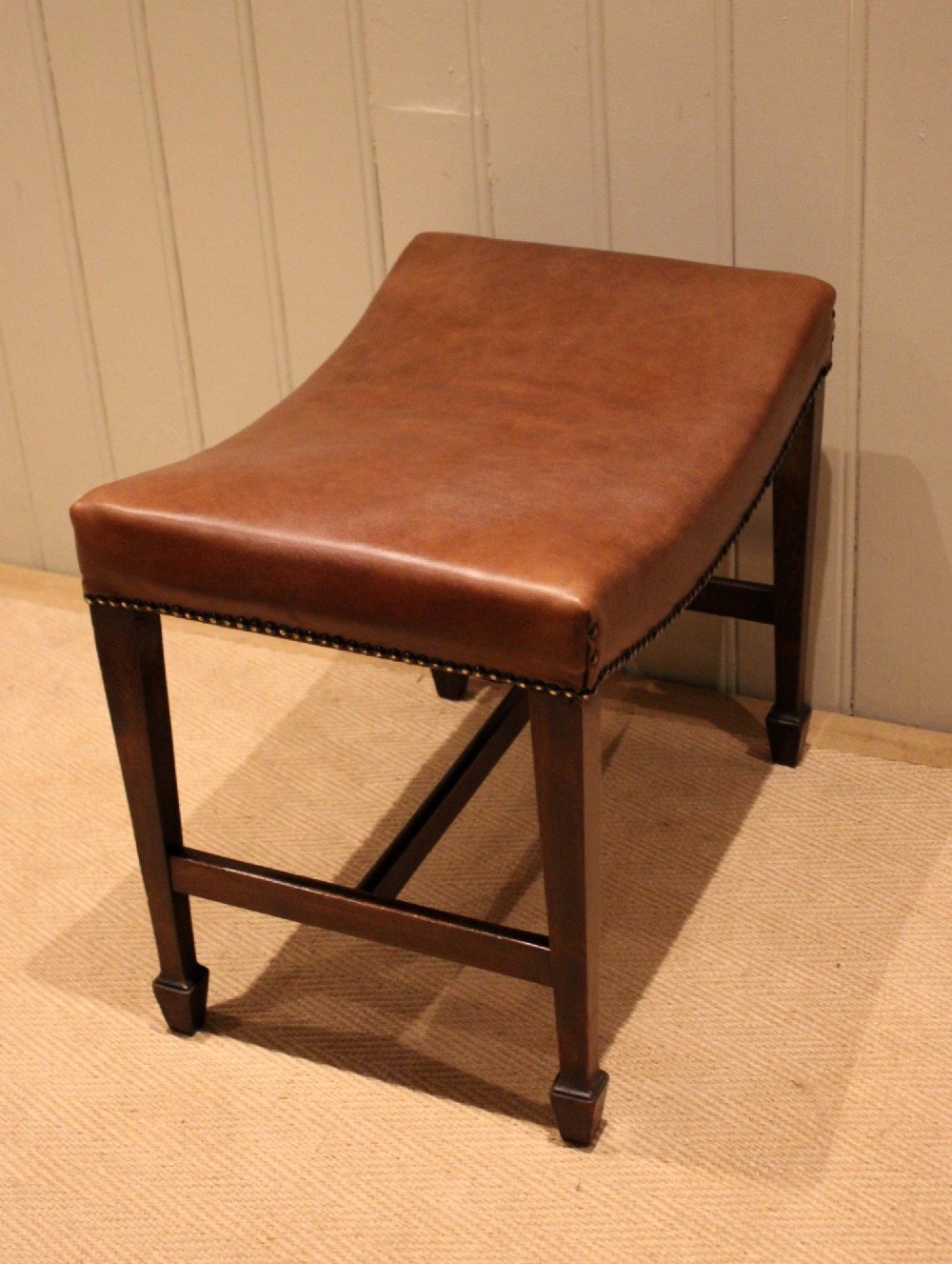 An Edwardian mahogany stool featuring a concave tan leather seat trimmed with brass studs and supported by square tapered legs with stretchers. View XII