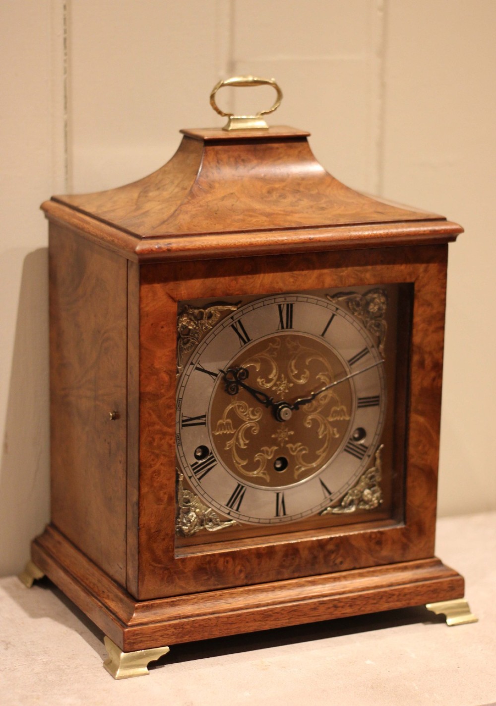 A burr walnut bracket clock featuring a caddy top with a brass carry handle, an ornate brass dial with a silvered chapter ring, and brass bracket feet. View II