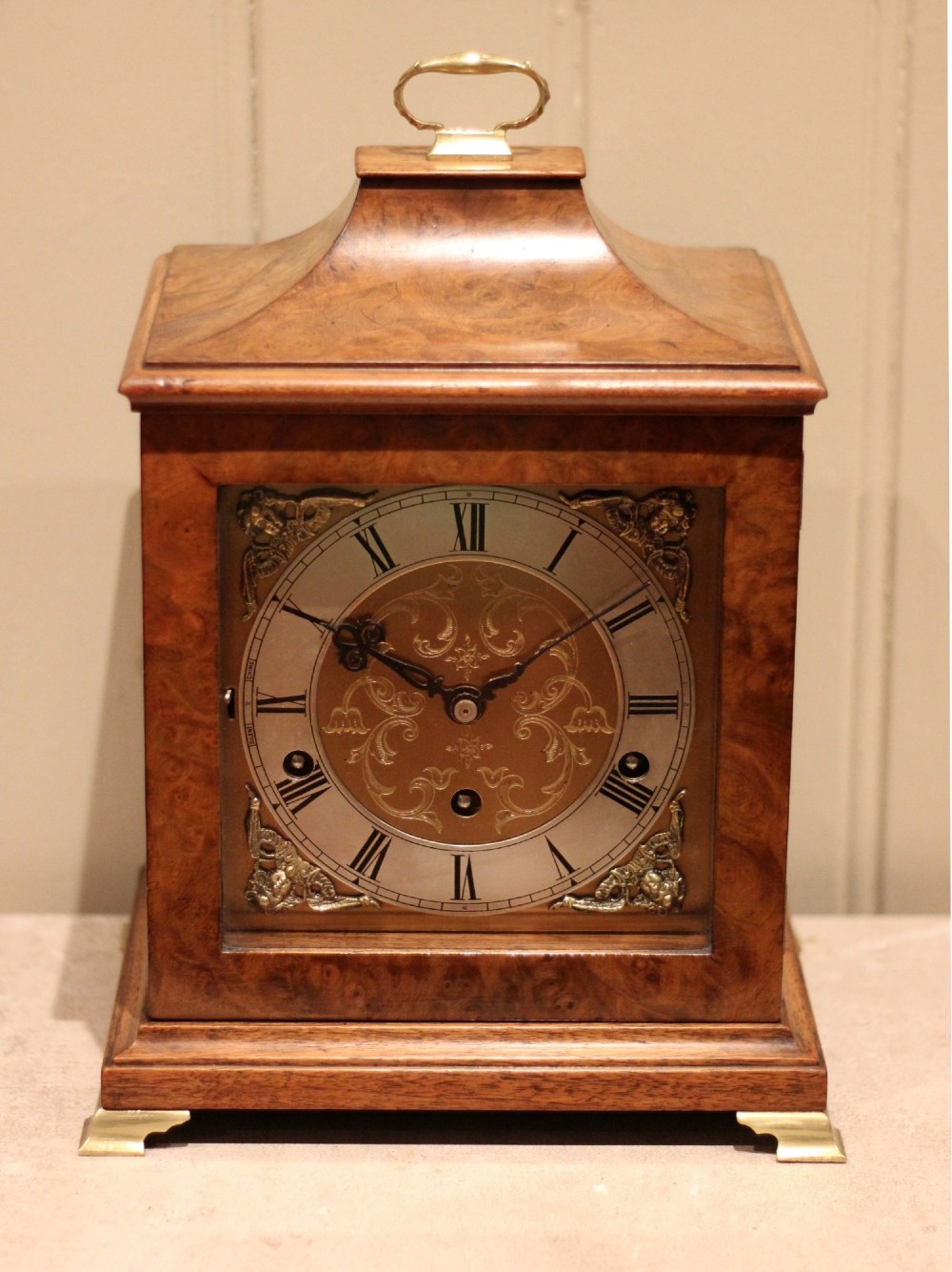 A burr walnut bracket clock featuring a caddy top with a brass carry handle, an ornate brass dial with a silvered chapter ring, and brass bracket feet. View V