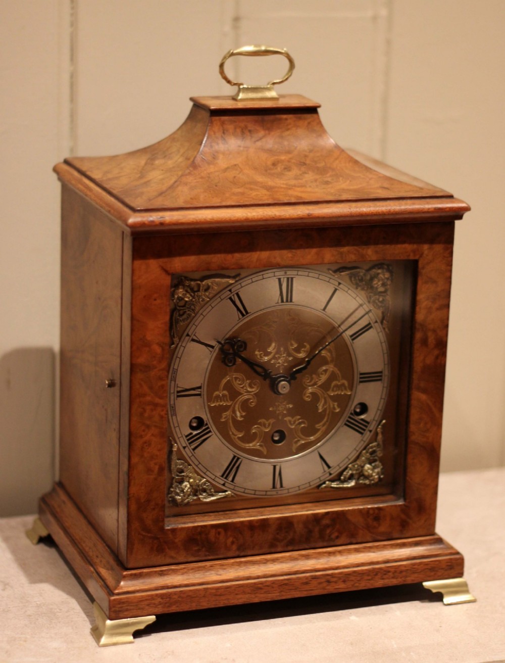 A burr walnut bracket clock featuring a caddy top with a brass carry handle, an ornate brass dial with a silvered chapter ring, and brass bracket feet. View VI