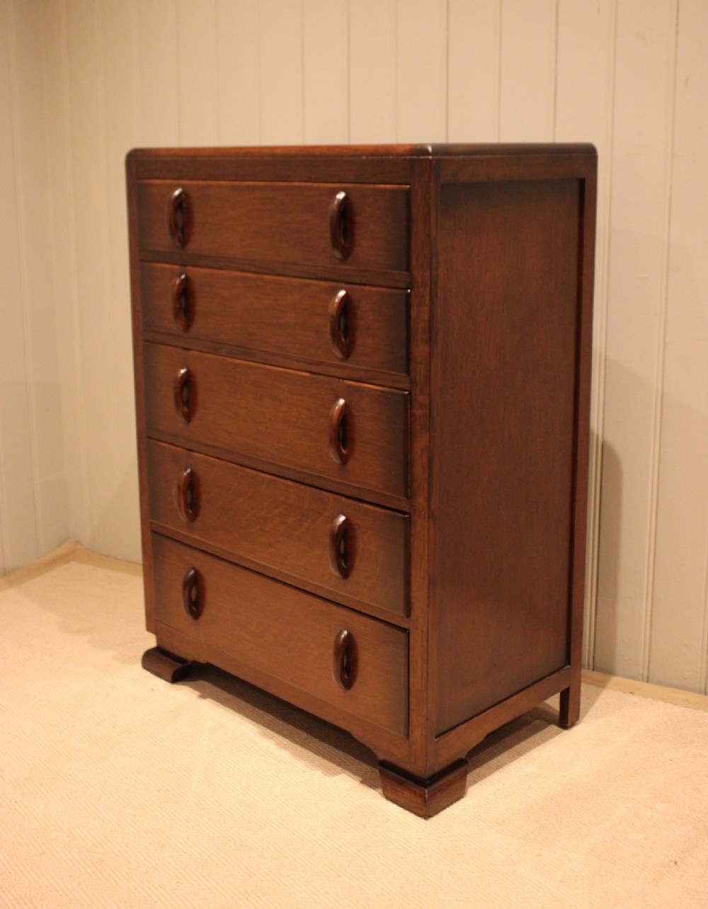 A dark brown oak Art Deco chest of drawers featuring five stacked drawers with matching wood handles and stepped feet. View X