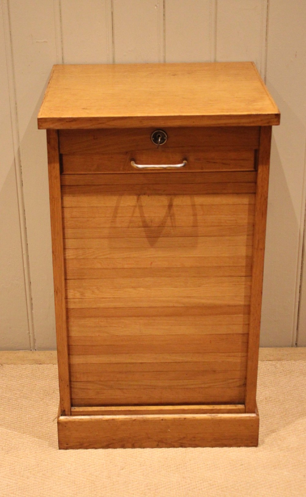 A small light oak filing cabinet featuring a slatted wooden tambour front with a metal handle and keyhole lock at the top. View V