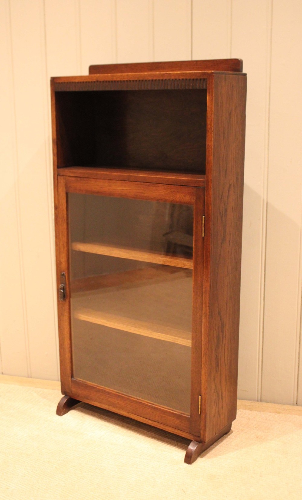 A medium oak freestanding bookcase featuring an open top compartment above a single glass-fronted door with internal shelving. View VI