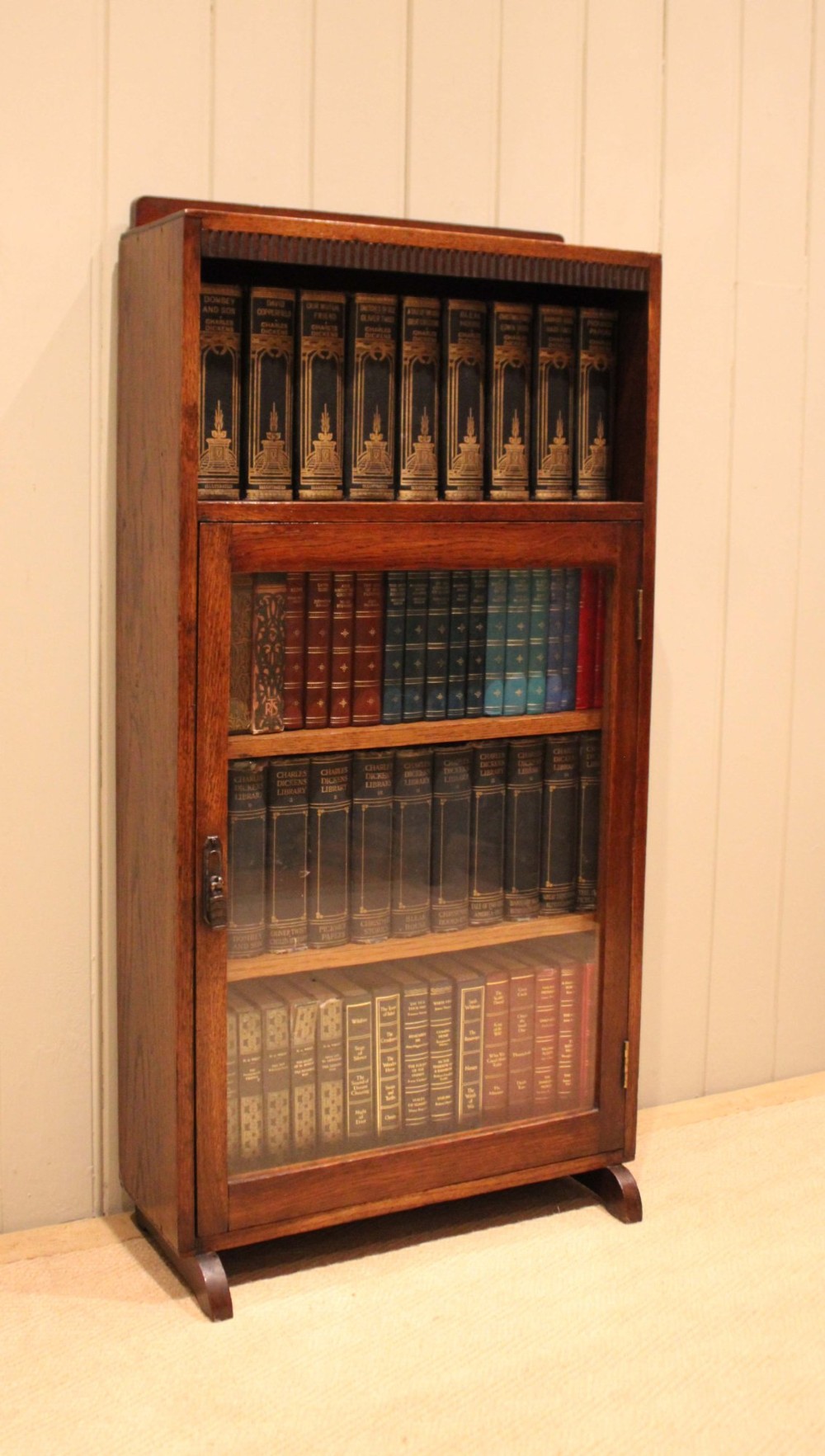 A medium oak freestanding bookcase featuring an open top compartment above a single glass-fronted door with internal shelving. View VII