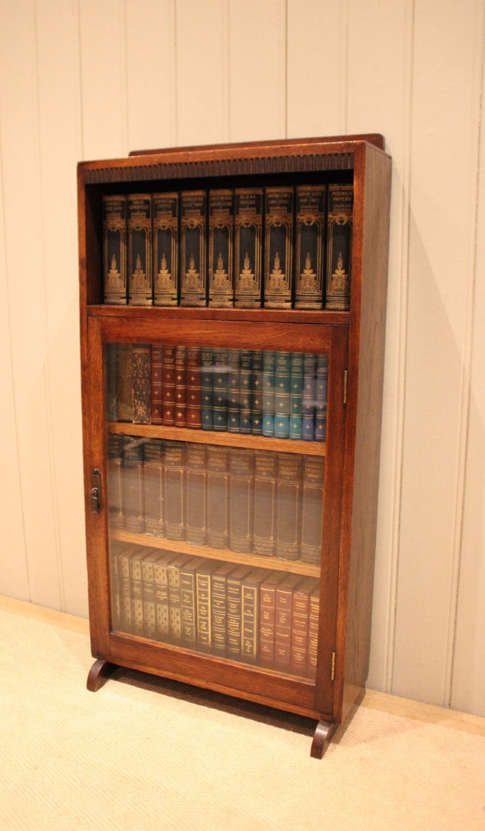 A medium oak freestanding bookcase featuring an open top compartment above a single glass-fronted door with internal shelving. View IX