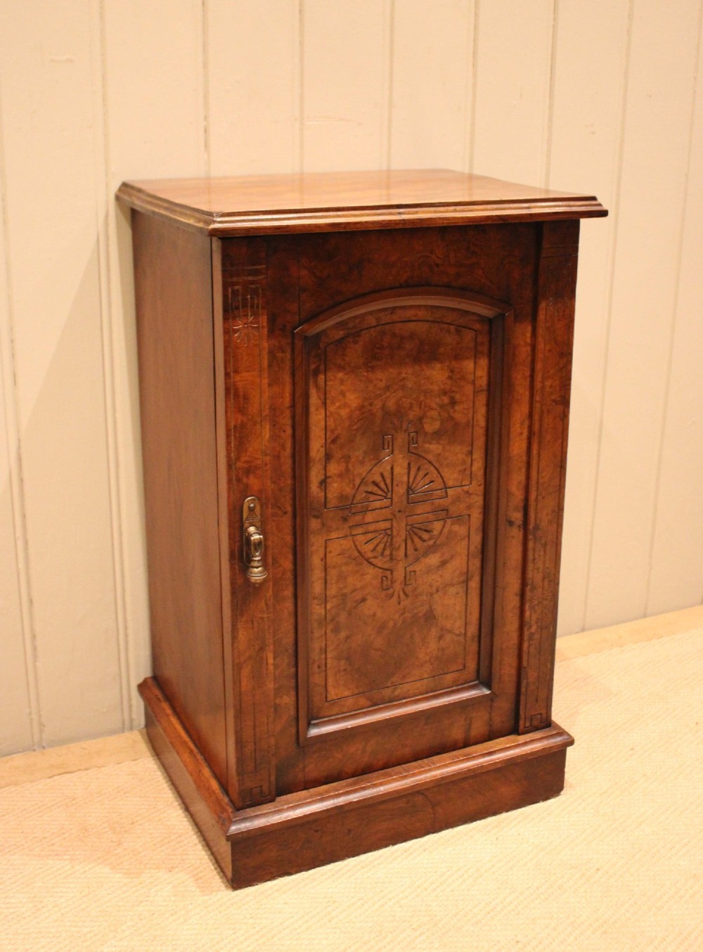 A late 19th-century walnut bedside cabinet featuring a single door with incised geometric detail, a brass drop handle, and a solid plinth base. View V