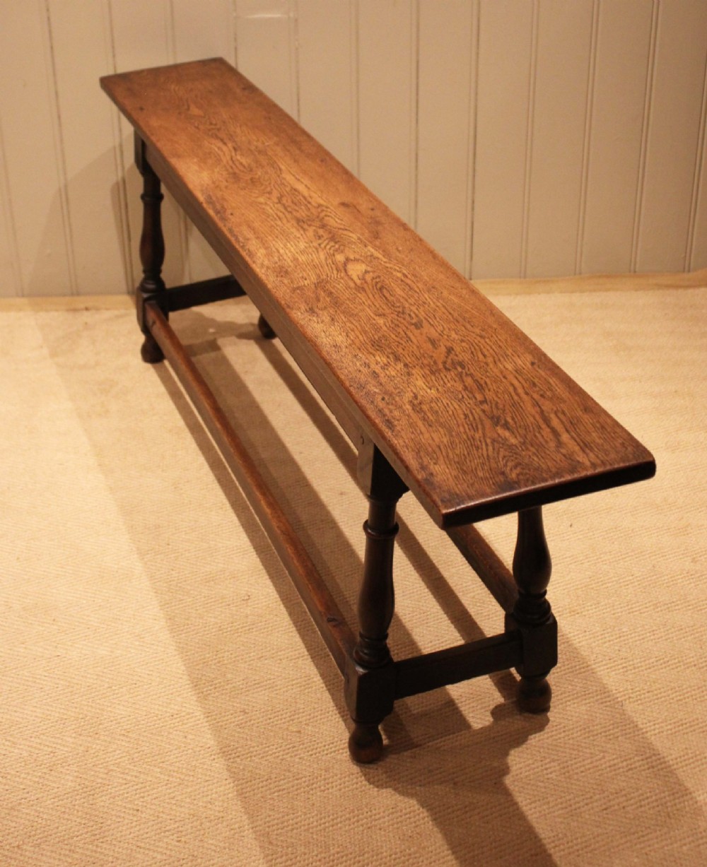 A long solid oak bench featuring a flat timber seat supported by four turned legs and a low rectangular stretcher base. View X