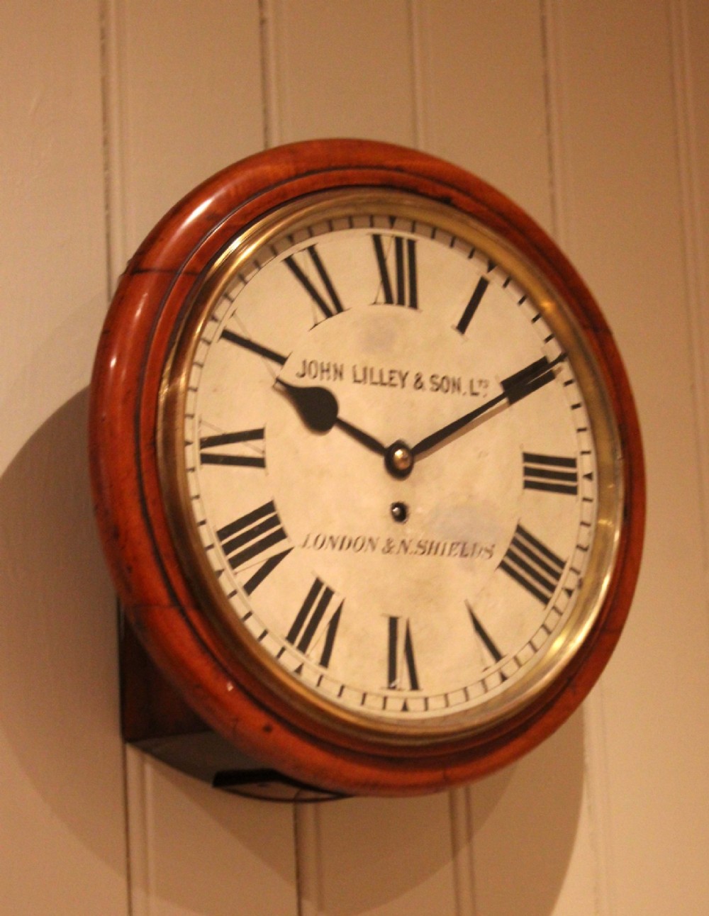 A circular late nineteenth-century Victorian wall clock featuring a turned mahogany frame, a polished brass bezel, and a cream dial with black Roman numerals. View V