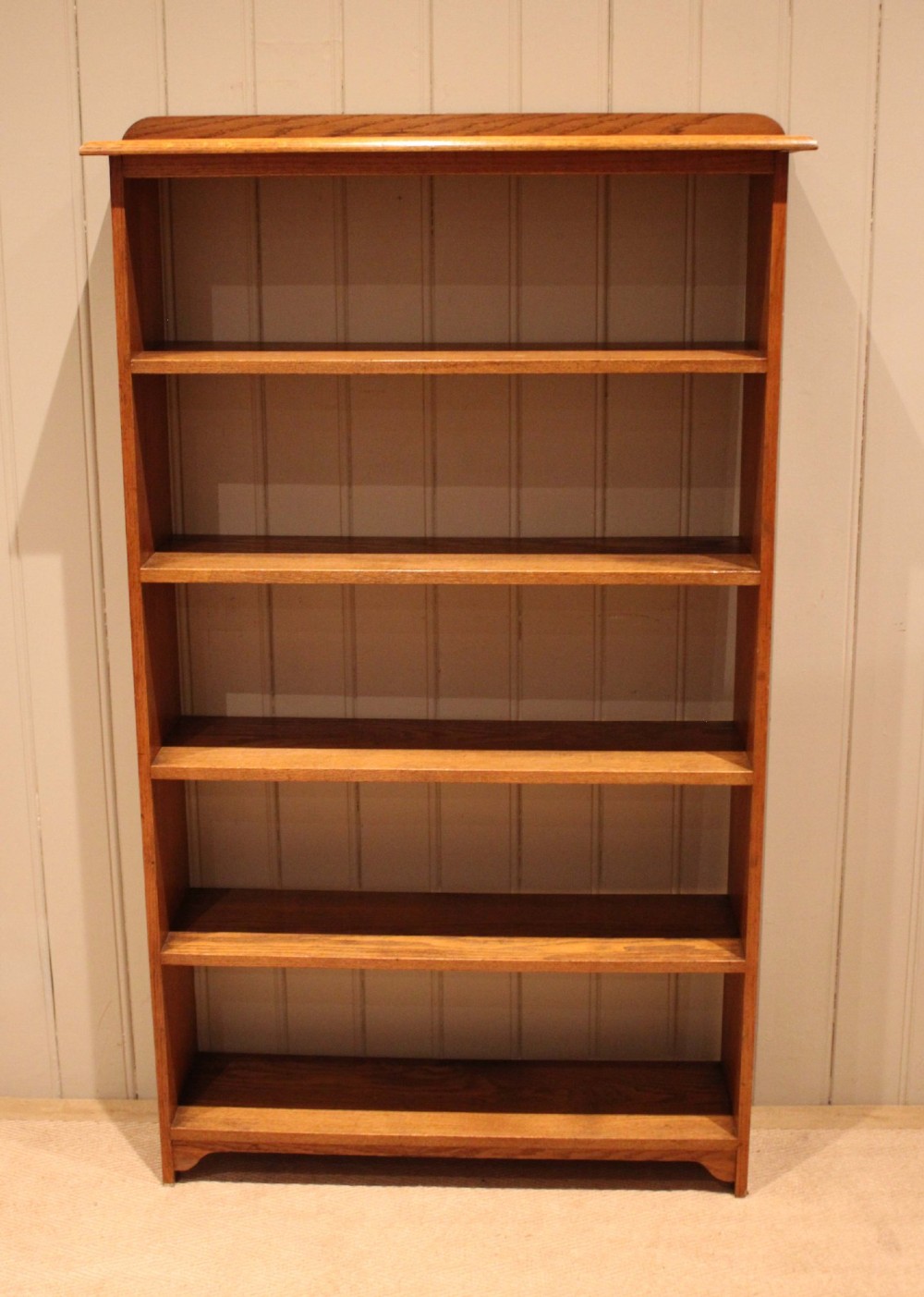 A tall, slender open oak bookcase with five tiers filled with multi-coloured hardback books, featuring a rounded top moulding and simple cut-out bracket feet. View II