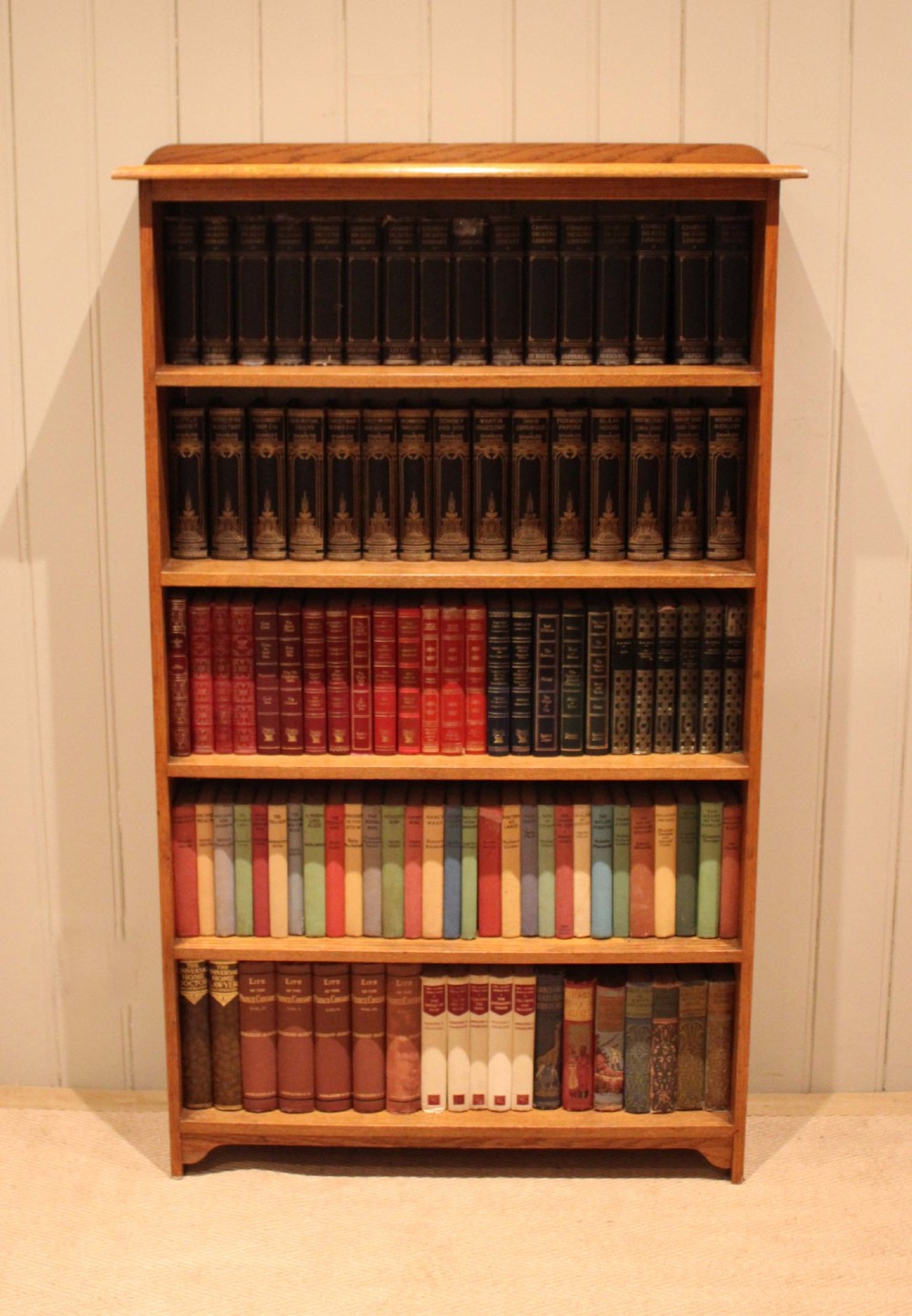 A tall, slender open oak bookcase with five tiers filled with multi-coloured hardback books, featuring a rounded top moulding and simple cut-out bracket feet. View III