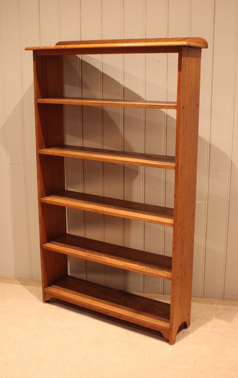 A tall, slender open oak bookcase with five tiers filled with multi-coloured hardback books, featuring a rounded top moulding and simple cut-out bracket feet. View IV