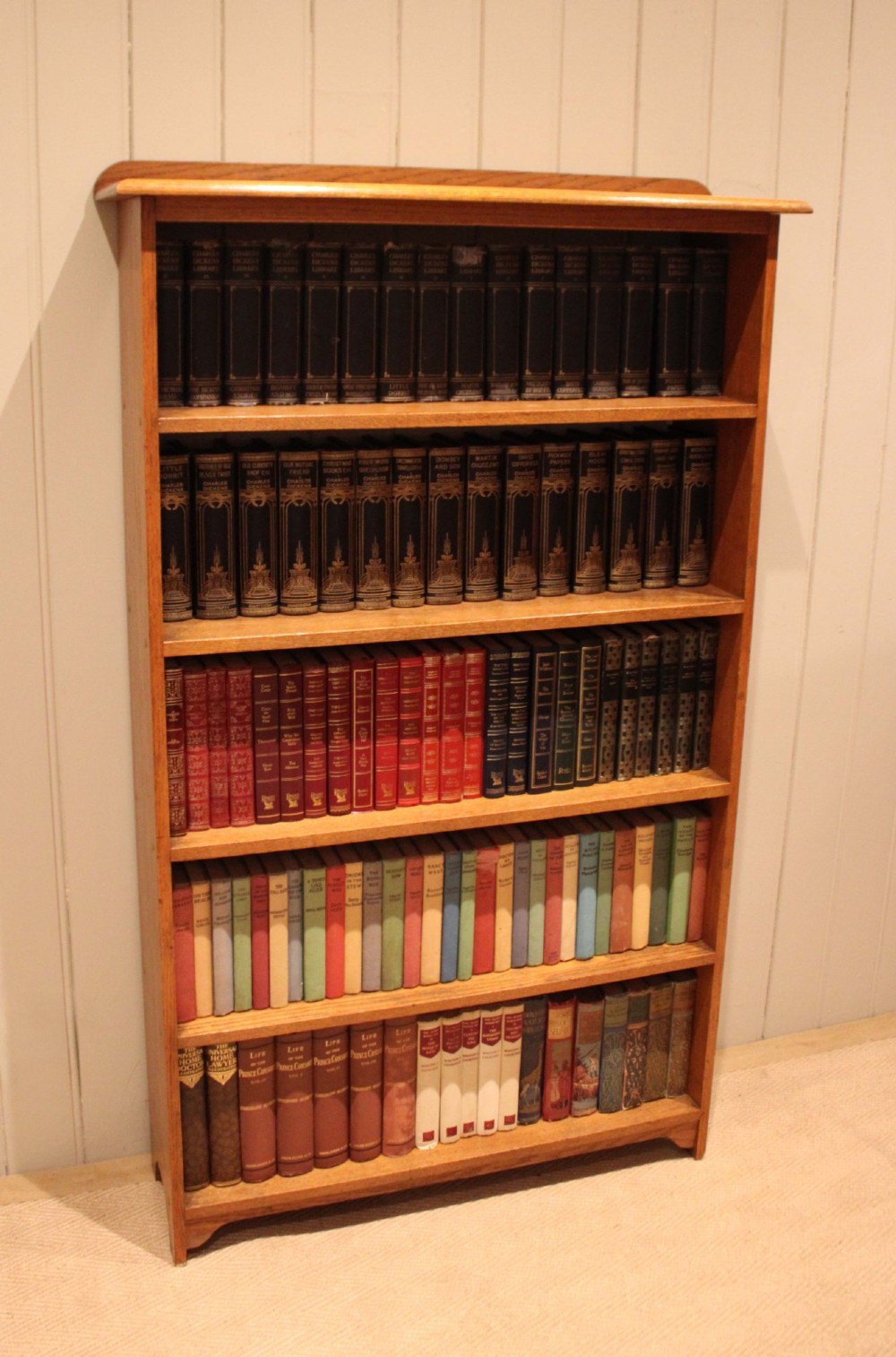 A tall, slender open oak bookcase with five tiers filled with multi-coloured hardback books, featuring a rounded top moulding and simple cut-out bracket feet. View V