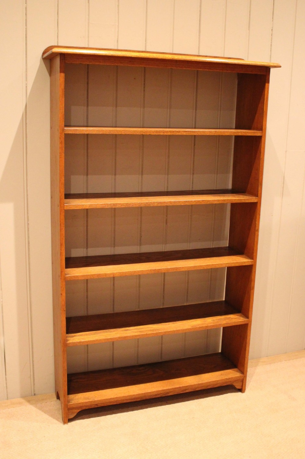A tall, slender open oak bookcase with five tiers filled with multi-coloured hardback books, featuring a rounded top moulding and simple cut-out bracket feet. View VI