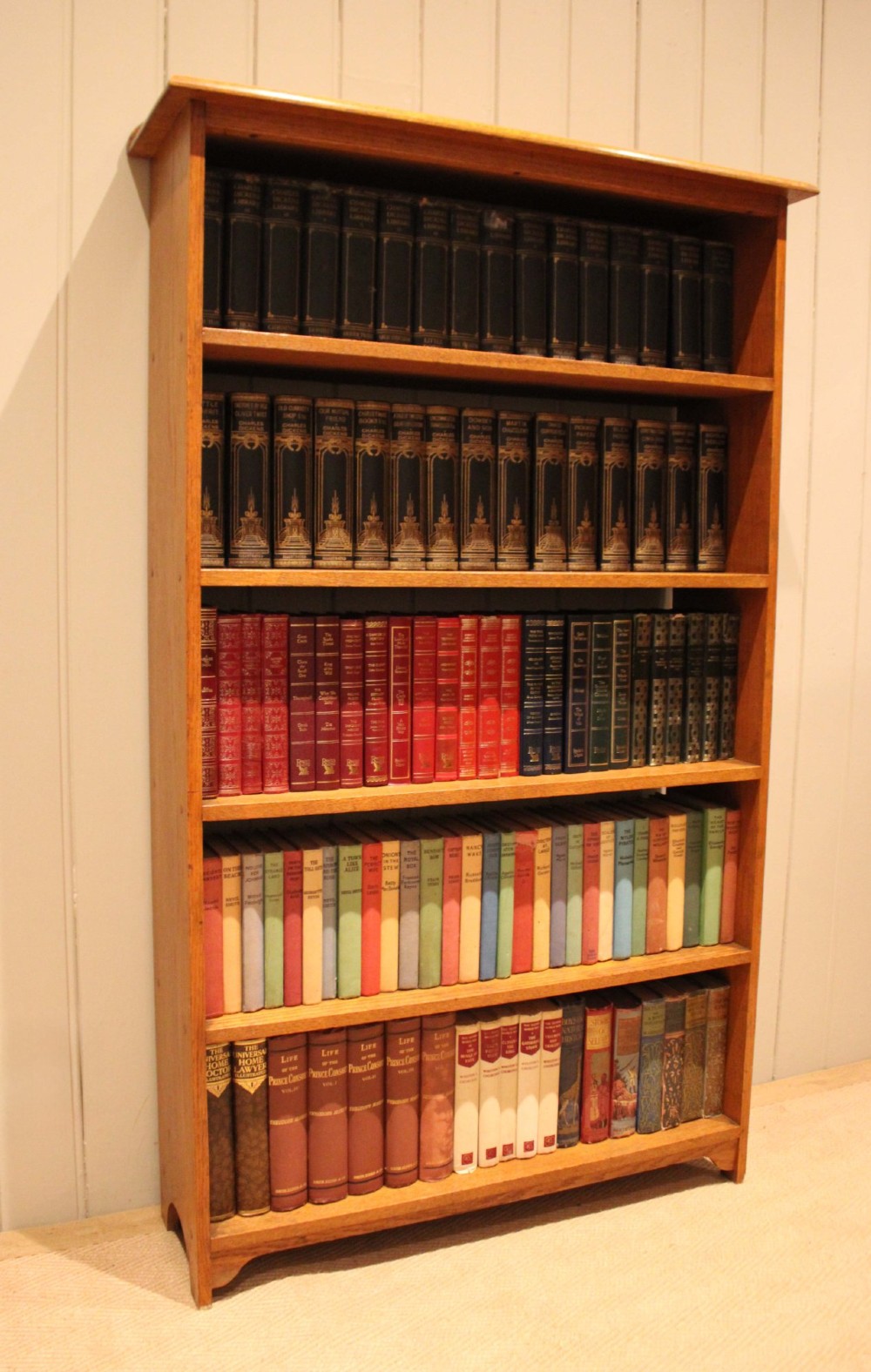 A tall, slender open oak bookcase with five tiers filled with multi-coloured hardback books, featuring a rounded top moulding and simple cut-out bracket feet. View VII