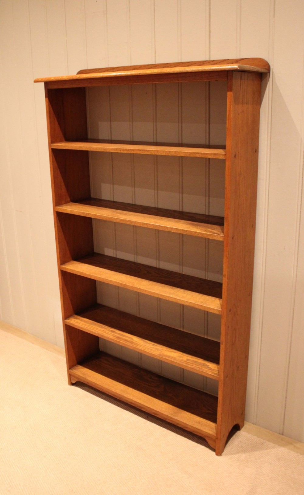 A tall, slender open oak bookcase with five tiers filled with multi-coloured hardback books, featuring a rounded top moulding and simple cut-out bracket feet. View VIII