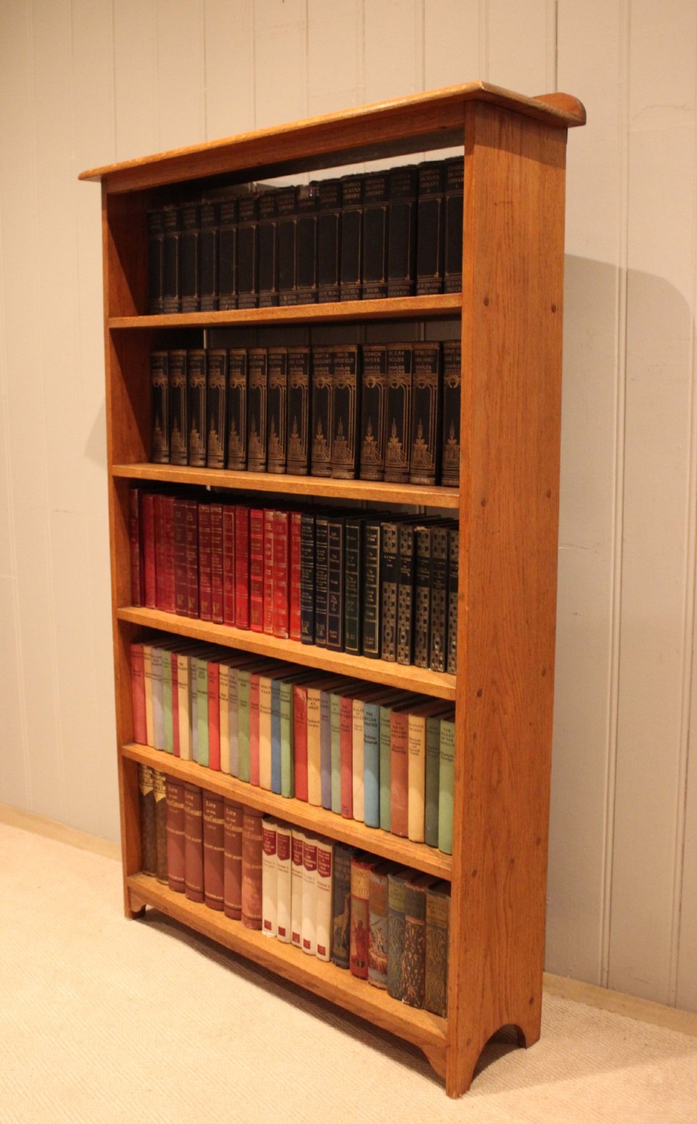 A tall, slender open oak bookcase with five tiers filled with multi-coloured hardback books, featuring a rounded top moulding and simple cut-out bracket feet. View IX