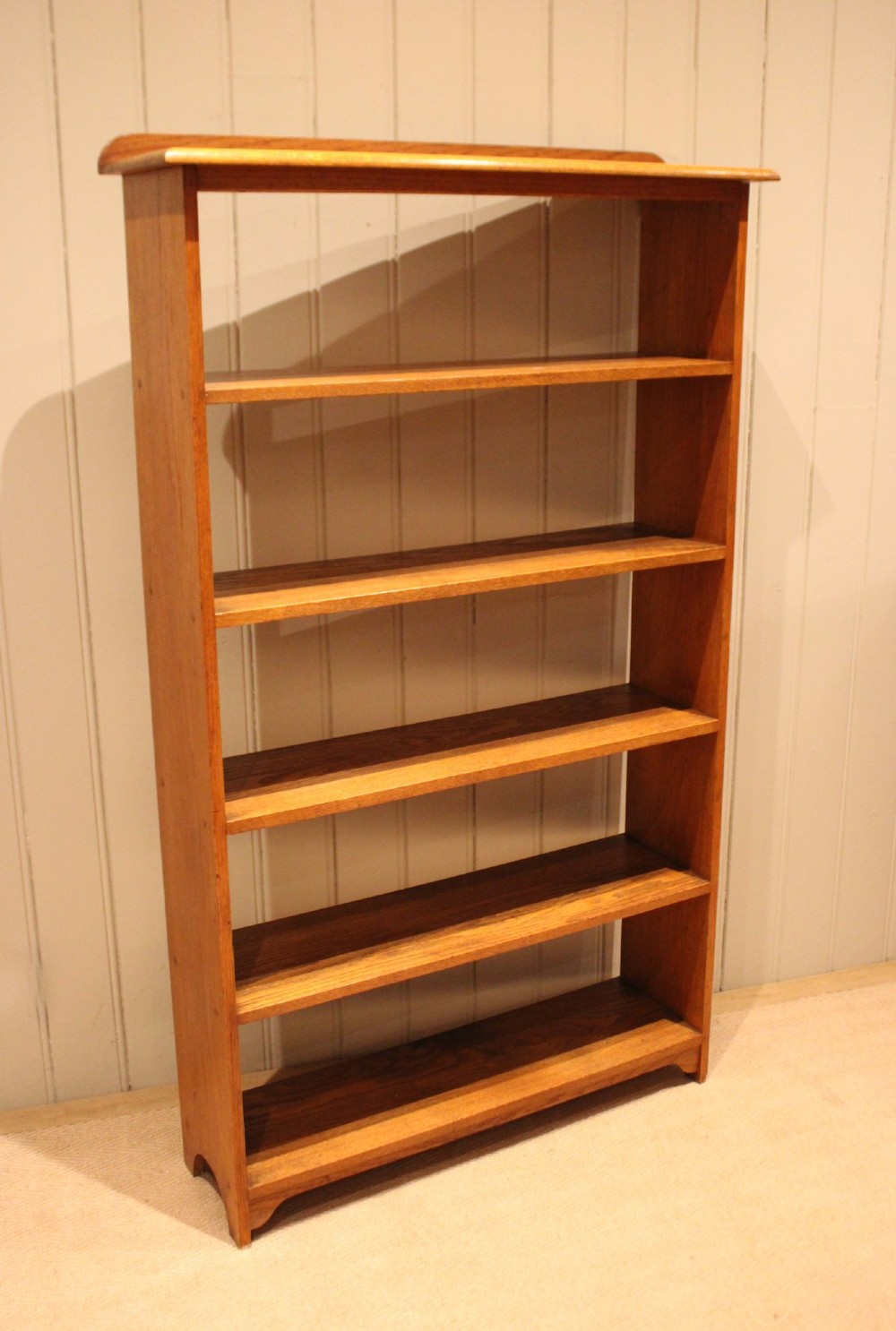 A tall, slender open oak bookcase with five tiers filled with multi-coloured hardback books, featuring a rounded top moulding and simple cut-out bracket feet. View X