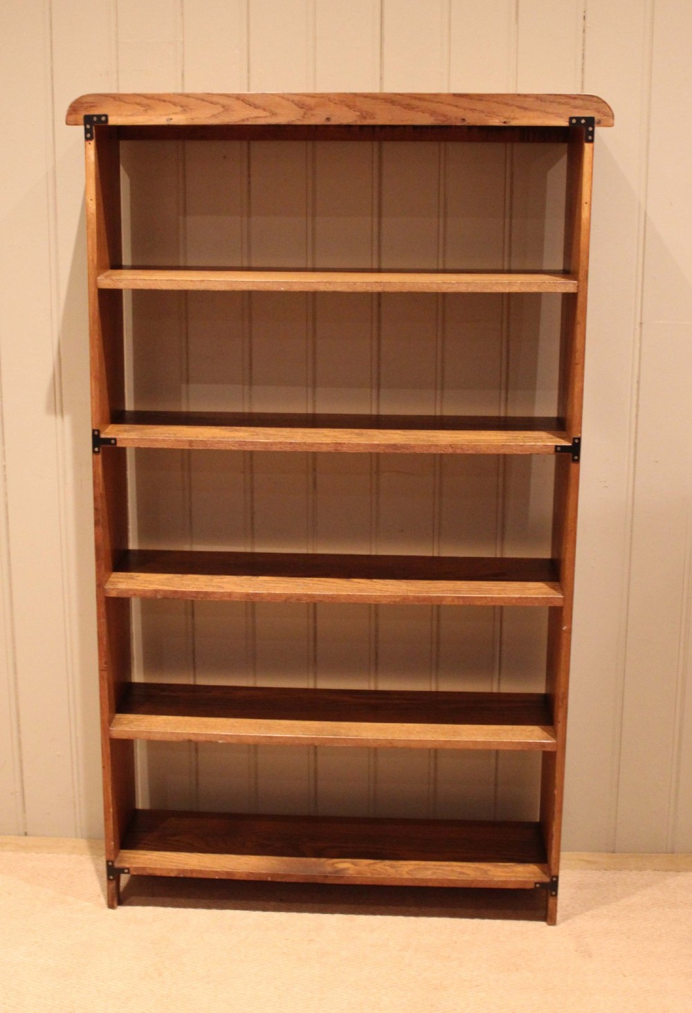 A tall, slender open oak bookcase with five tiers filled with multi-coloured hardback books, featuring a rounded top moulding and simple cut-out bracket feet. View XII