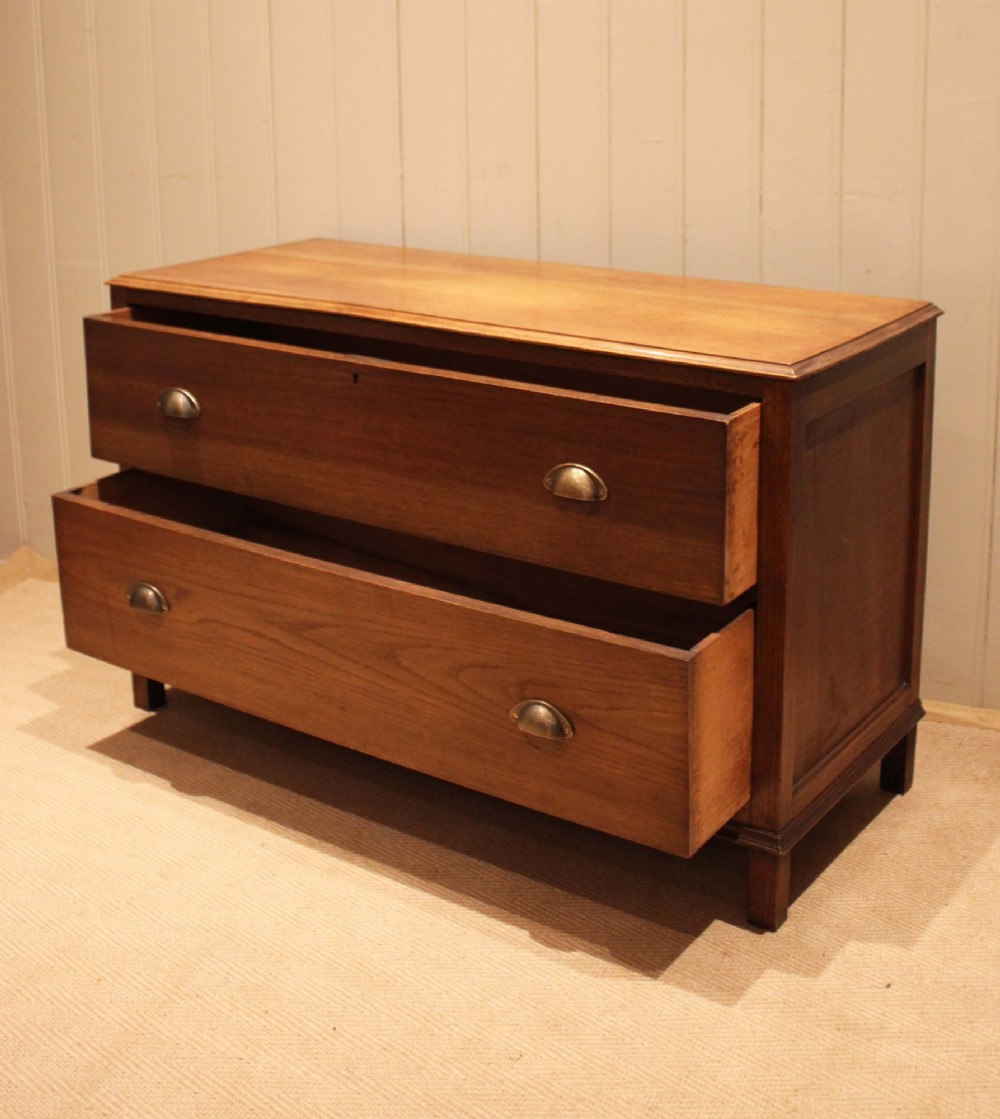 A wide low oak chest of drawers featuring two deep drawers with brass shell cup handles and panelled side construction. View VI
