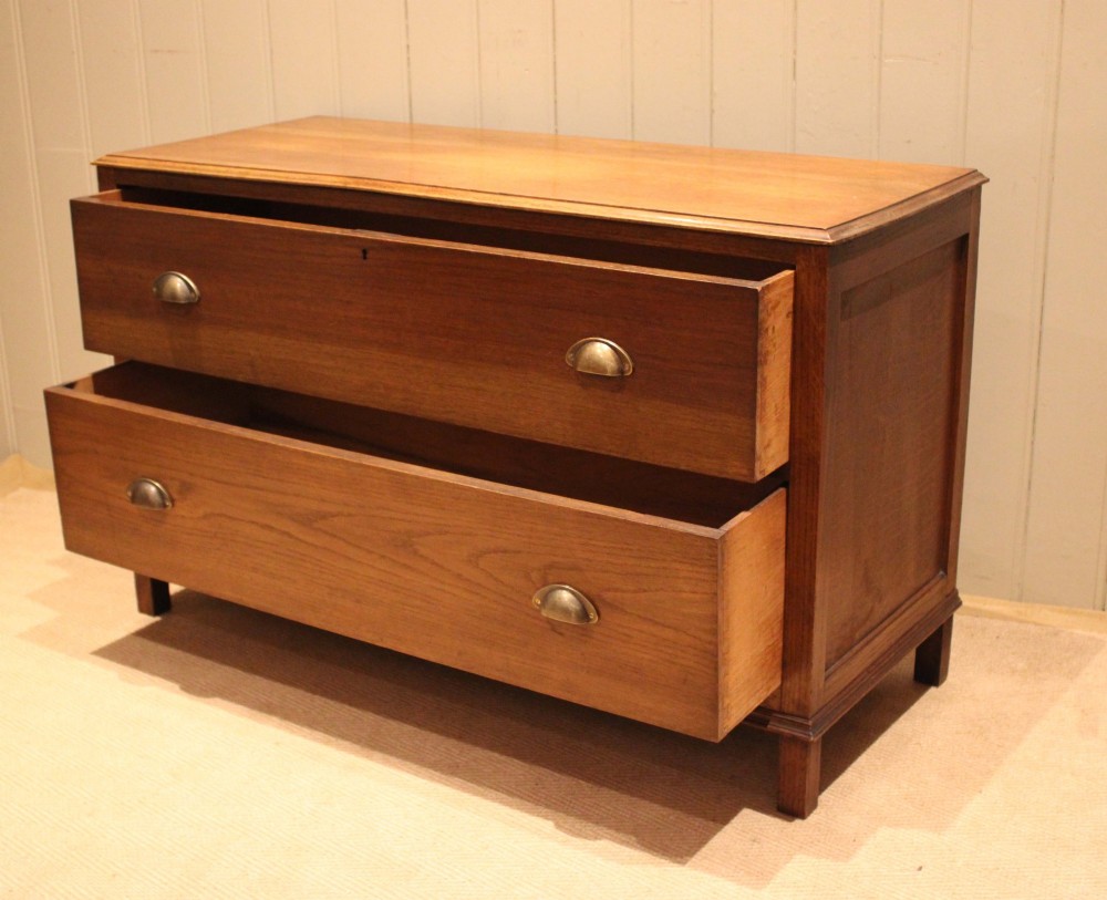 A wide low oak chest of drawers featuring two deep drawers with brass shell cup handles and panelled side construction. View XI