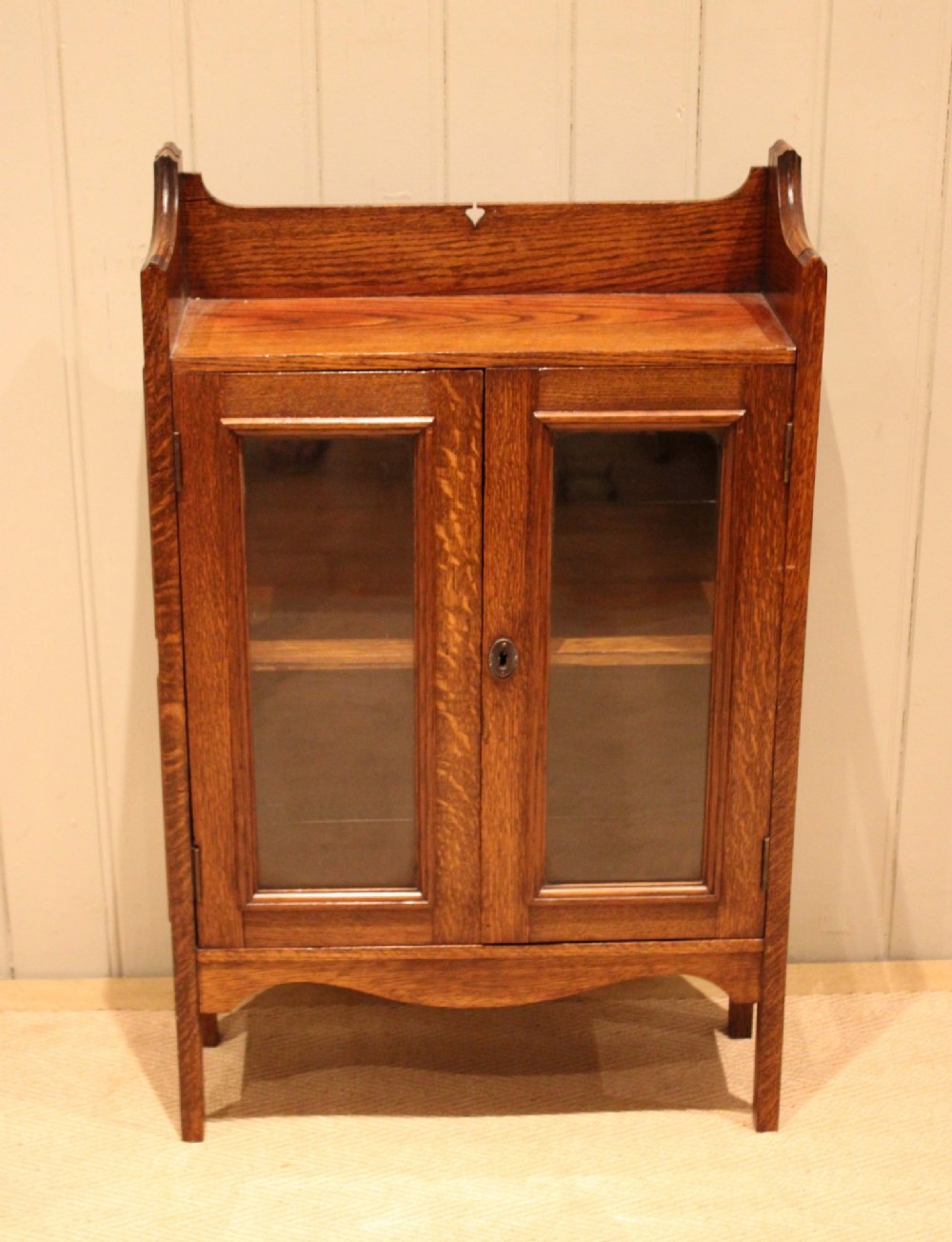 A small Arts and Crafts medium oak glazed bookcase featuring a gallery top with a pierced heart motif, two glazed doors, glazed side panels, and square legs. View II