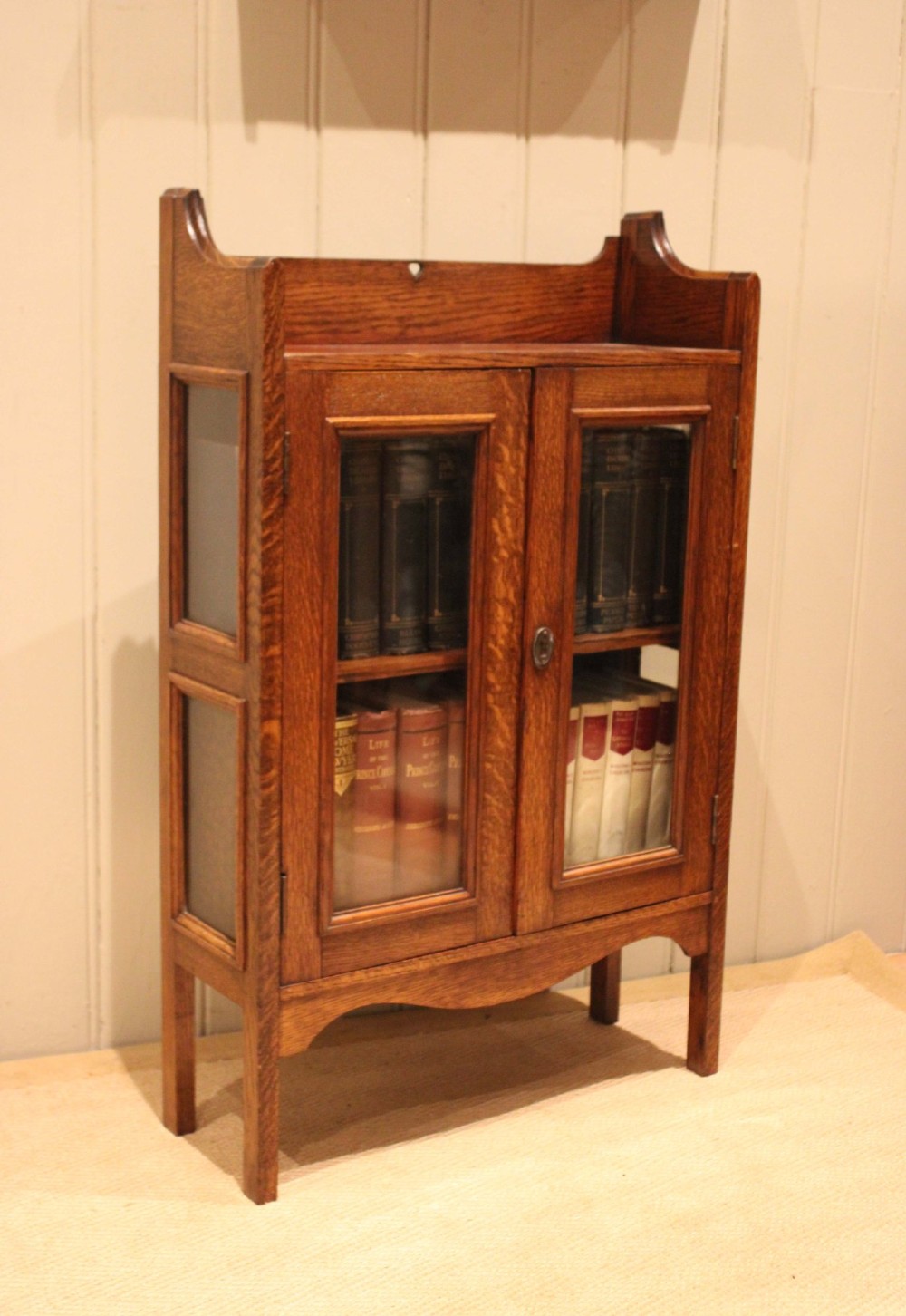 A small Arts and Crafts medium oak glazed bookcase featuring a gallery top with a pierced heart motif, two glazed doors, glazed side panels, and square legs. View V