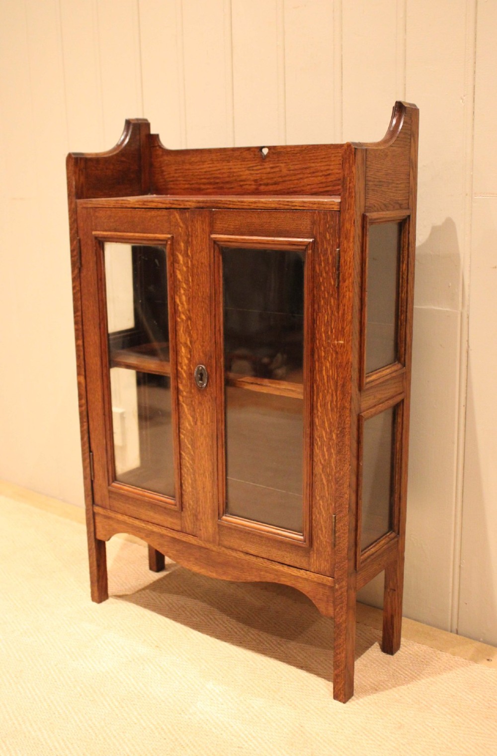 A small Arts and Crafts medium oak glazed bookcase featuring a gallery top with a pierced heart motif, two glazed doors, glazed side panels, and square legs. View VI