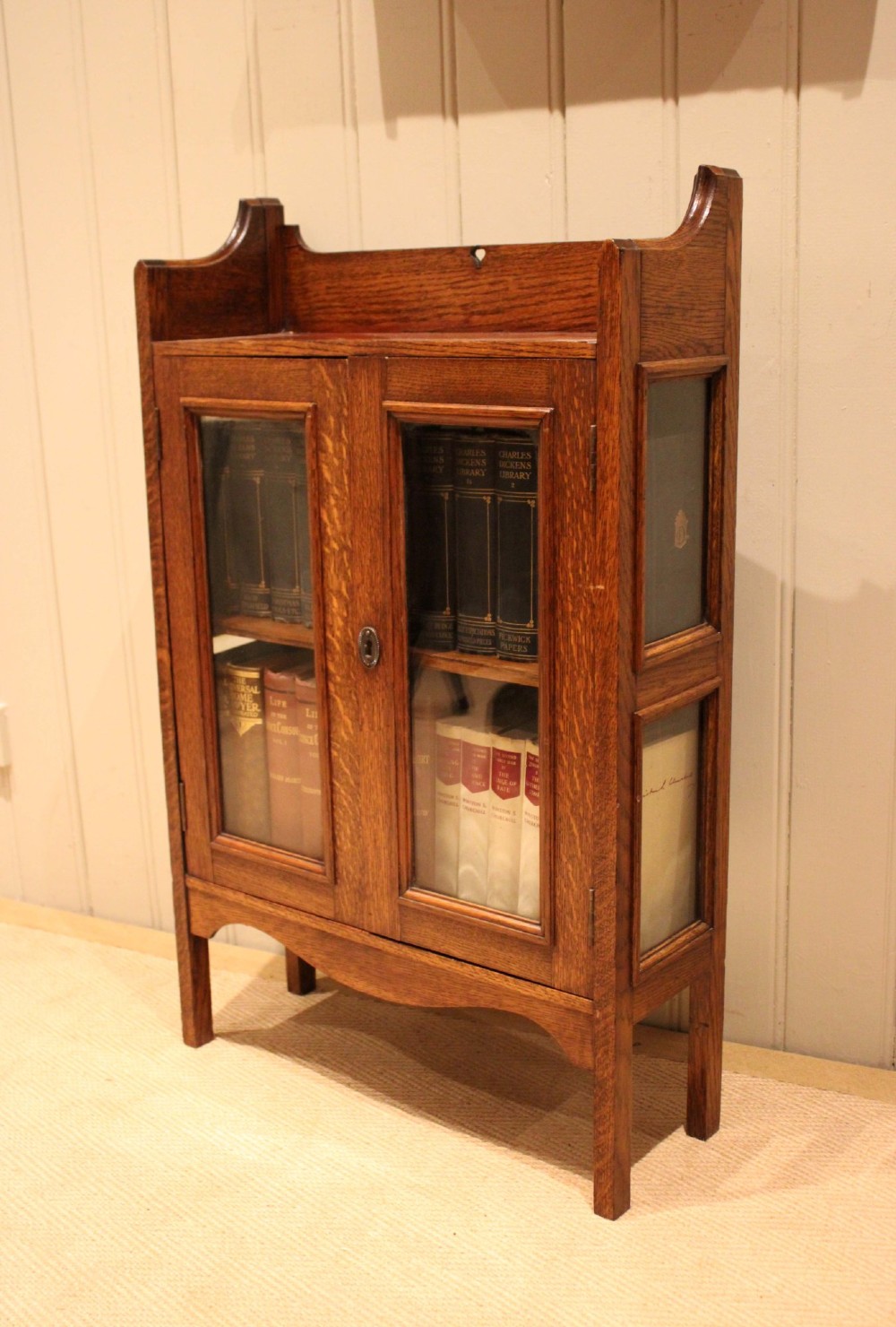 A small Arts and Crafts medium oak glazed bookcase featuring a gallery top with a pierced heart motif, two glazed doors, glazed side panels, and square legs. View VII