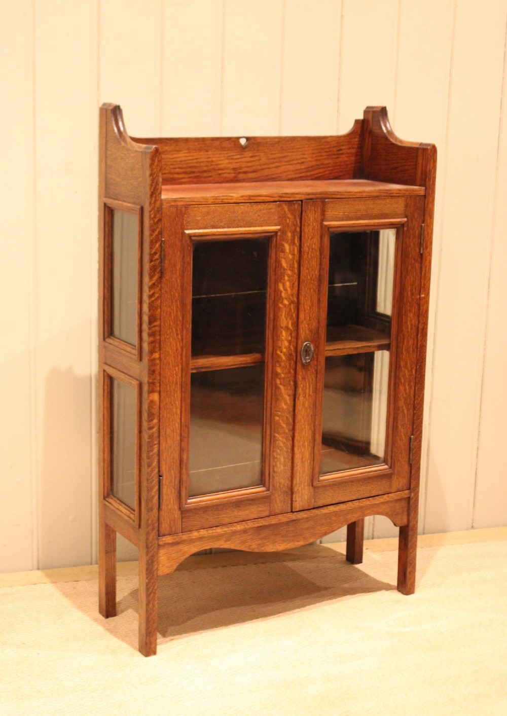 A small Arts and Crafts medium oak glazed bookcase featuring a gallery top with a pierced heart motif, two glazed doors, glazed side panels, and square legs. View VIII