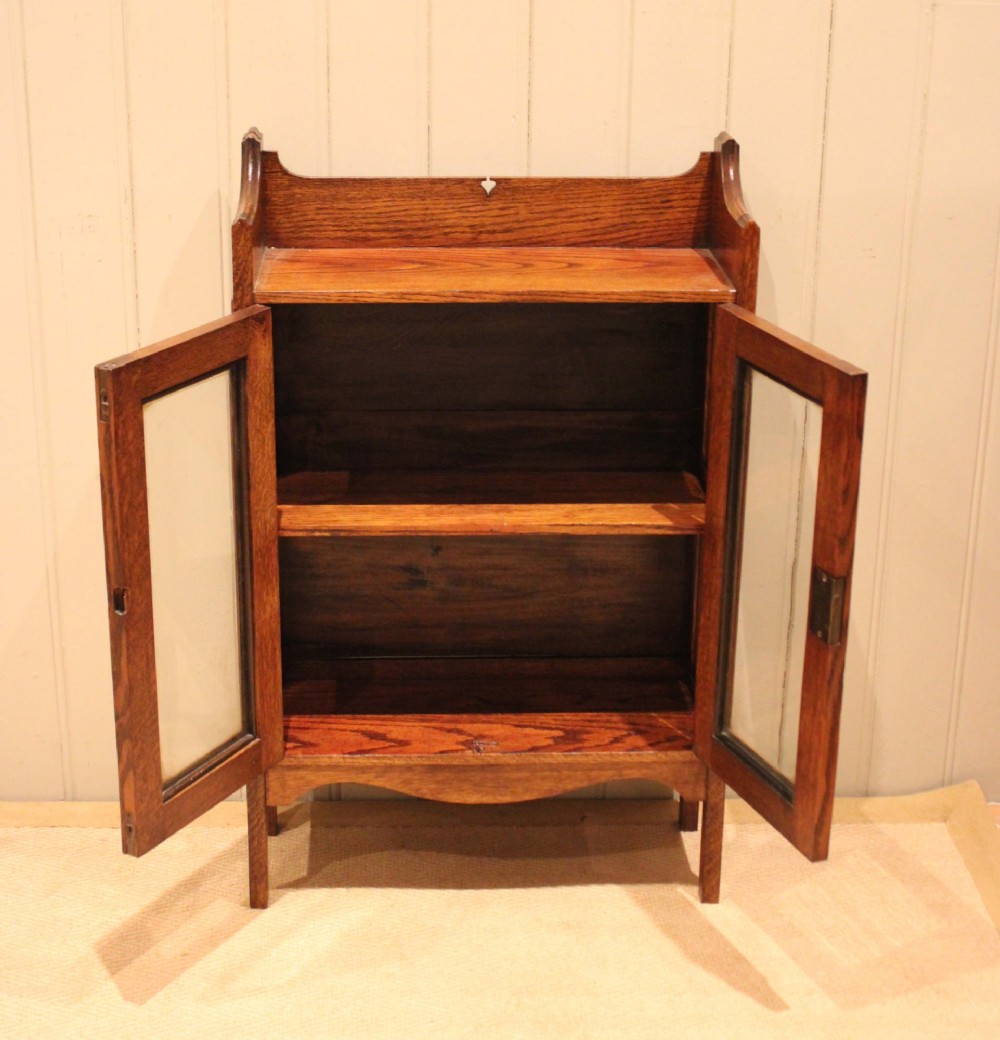 A small Arts and Crafts medium oak glazed bookcase featuring a gallery top with a pierced heart motif, two glazed doors, glazed side panels, and square legs. View X