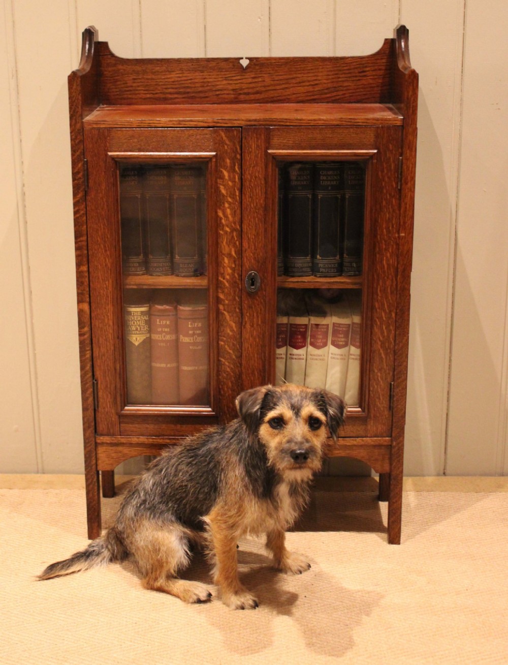 A small Arts and Crafts medium oak glazed bookcase featuring a gallery top with a pierced heart motif, two glazed doors, glazed side panels, and square legs. View XI