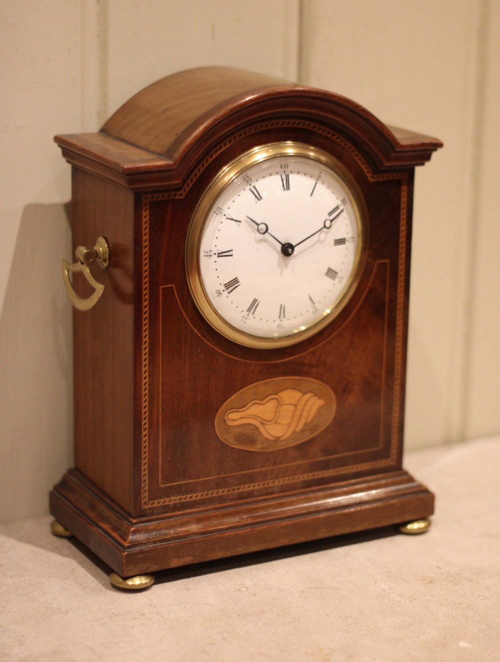 An Edwardian arched mahogany mantel clock featuring a shell motif inlay, a white dial with Roman numerals, brass side handles, and brass ball feet. View II