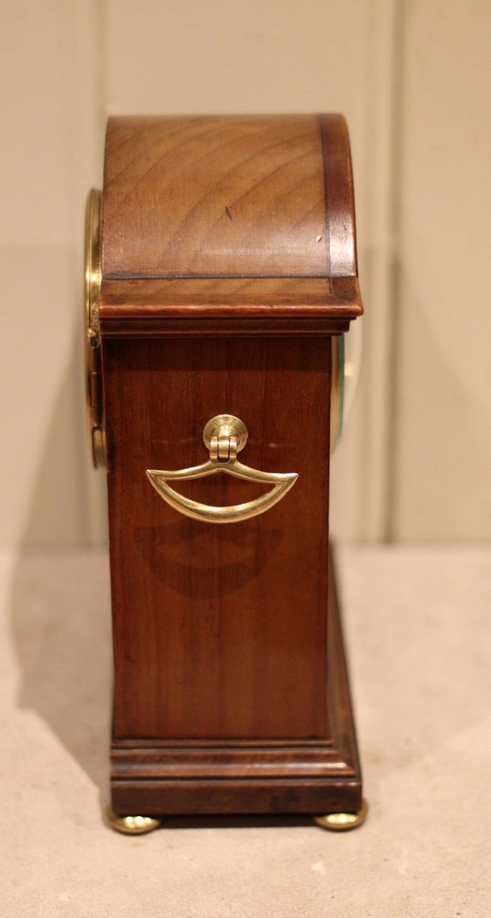 An Edwardian arched mahogany mantel clock featuring a shell motif inlay, a white dial with Roman numerals, brass side handles, and brass ball feet. View V