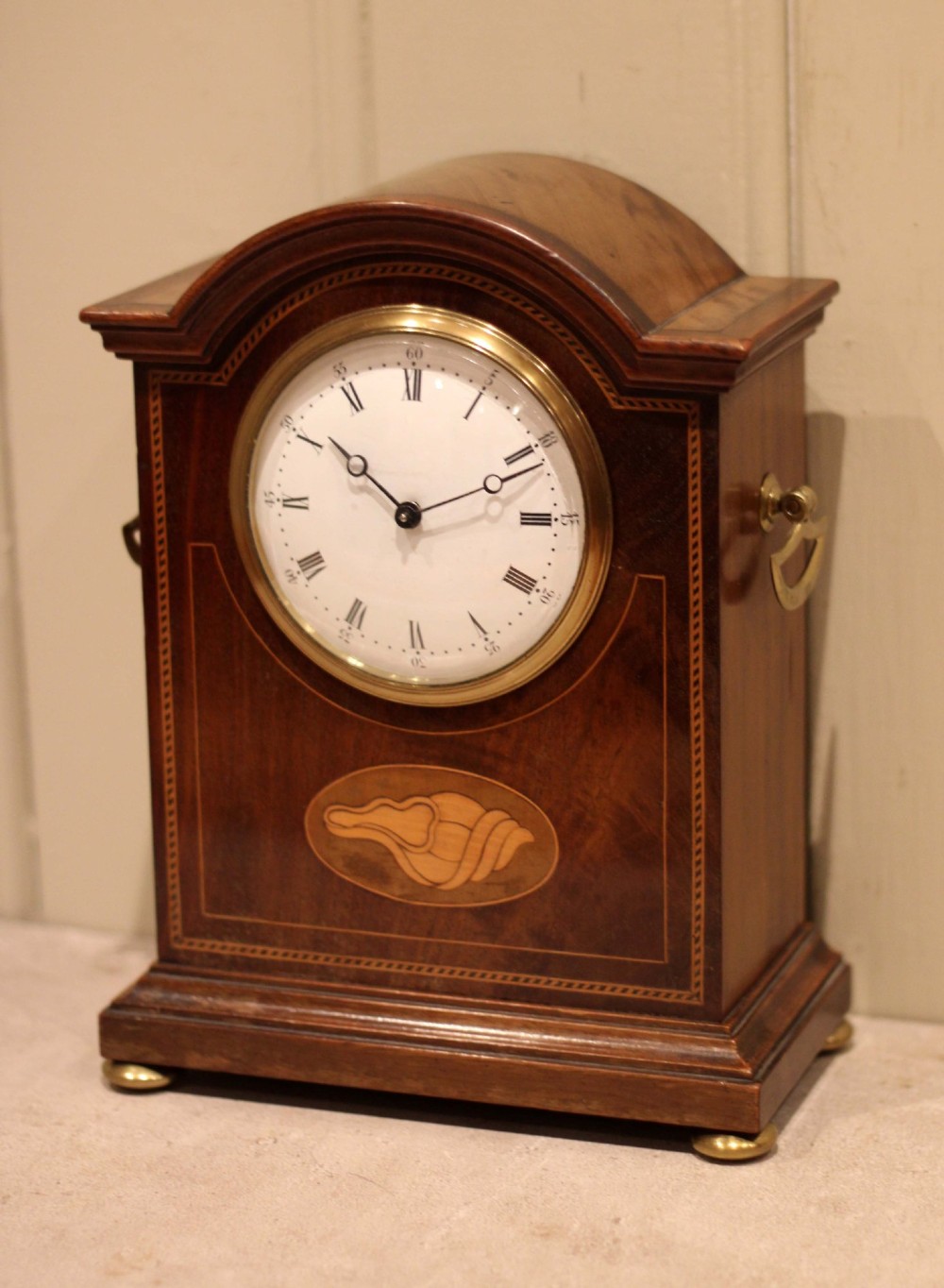 An Edwardian arched mahogany mantel clock featuring a shell motif inlay, a white dial with Roman numerals, brass side handles, and brass ball feet. View VI