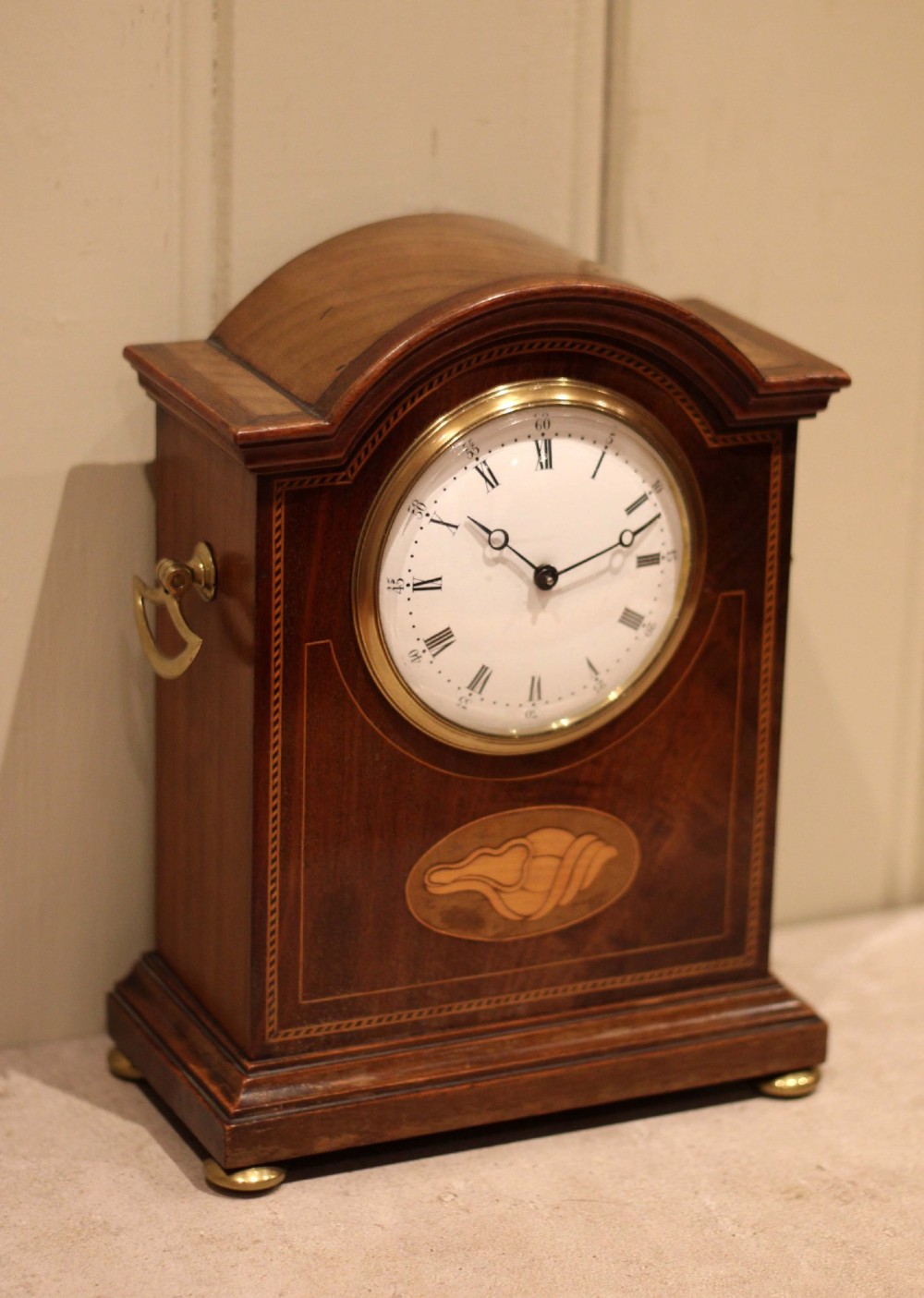 An Edwardian arched mahogany mantel clock featuring a shell motif inlay, a white dial with Roman numerals, brass side handles, and brass ball feet. View VII