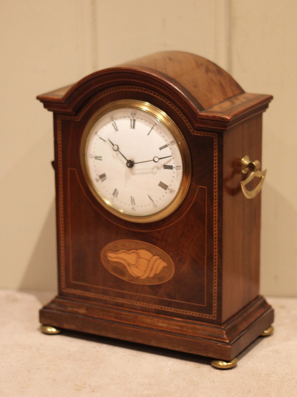 An Edwardian arched mahogany mantel clock featuring a shell motif inlay, a white dial with Roman numerals, brass side handles, and brass ball feet. View VIII