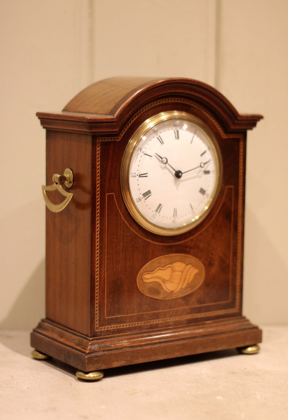 An Edwardian arched mahogany mantel clock featuring a shell motif inlay, a white dial with Roman numerals, brass side handles, and brass ball feet. View IX