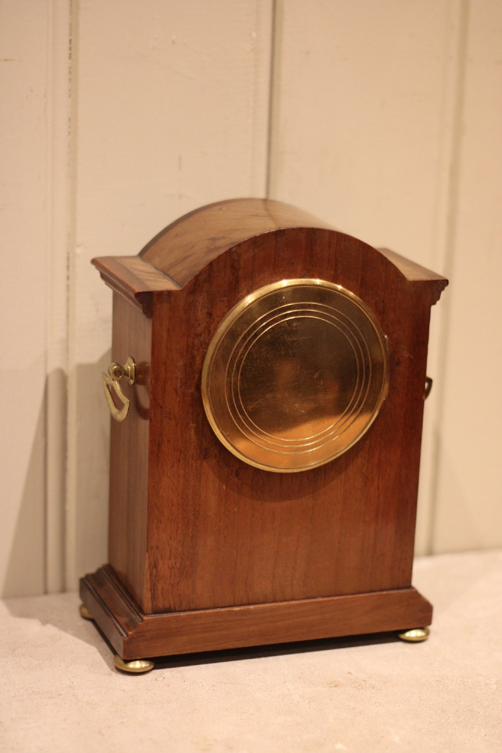 An Edwardian arched mahogany mantel clock featuring a shell motif inlay, a white dial with Roman numerals, brass side handles, and brass ball feet. View XI