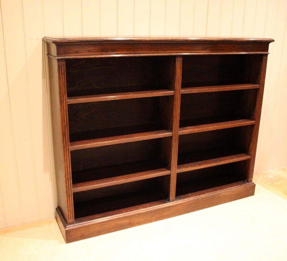 A double-bay open oak library bookcase featuring fluted pilasters, multiple horizontal shelves, a moulded top edge, and a solid plinth base. View X