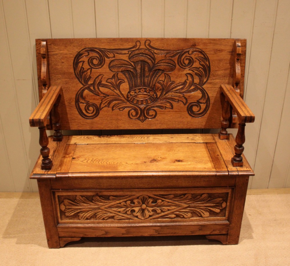 A carved solid oak monks bench featuring a decorative back panel that tilts forward to form a table surface, turned arm supports, and a lift-up storage seat. View II
